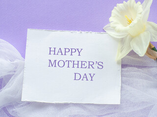 Daffodils flower and card with Happy Mothers day on lilac background. Mothers Day greeting card. Sheet of paper with text and narcissus on purple background. Congratulations note flat lay.