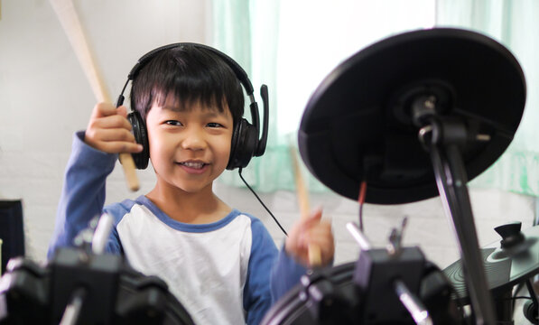 Asian child boy hold drumstick to play electronic drum with headphone and happy smiling face at home. Concept of music learning, happy relaxing time, freedom, little kid musician.
