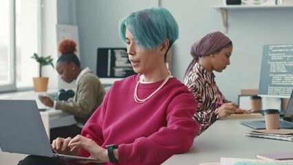 Blue haired young Asian male programmer using laptop while working in contemporary office with diverse female colleagues - Powered by Adobe