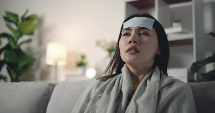 Footage of Sick young Asian woman in warm blanket shiver tremble with cold with cooler gel on her head on sofa at living room. People, health care and lifestyle concepts.