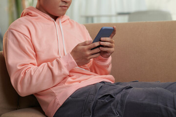 Serious young man sitting on couch at home and texting friends