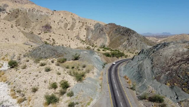 Aerial drone forward moving shot flying over car and truck driving along the winding RCD Road surrounded by arid vegetation in Balochistan on a sunny day.