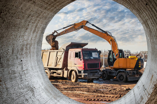 A wheeled excavator loads a dump truck with soil and sand. An excavator with a high-raised bucket against a cloudy sky View from the trench. Removal of soil from a construction site or quarry.
