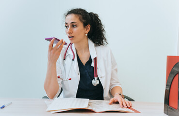 Brunette female Spanish doctor sits at desk records audio recommendation using phone at hospital....