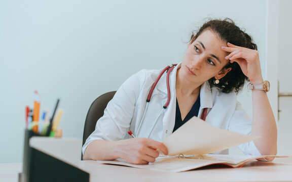 Tired female doctor sitting at desk thoughtfully looks away holds diagnosis of patient. Upset medical employee at workplace. Exhausted nurse leans on head with pensive face expression.