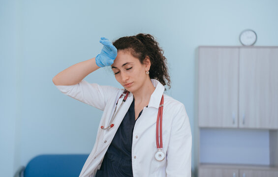 Exhausted female doctor in medical clothes touches temple eyes closed, tired after hard work. Young nurse tired stands at medical office with hands in  medical gloves.