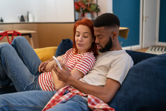 Young millennial diverse couple man and woman resting at home with mobile phone, hugging and surfing internet while relaxing together on sofa in living room. Family bonding and smartphone