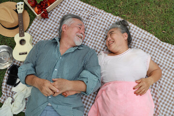 Top view happy asian senior man and woman lying on blanket and having fun on picnic together in garden outdoor. Lover couple eating food and embracing at the park. Happiness marriage lifestyle concept