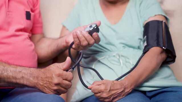 Close up shot of indian senior man checking bp or blood pressure of wife by using sphygmomanometer at home - concept of routine health checkup, medical care and togetherness.