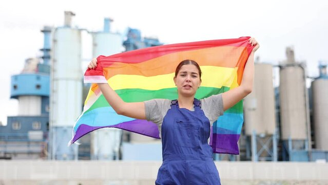 Against backdrop of factory located outside city,balanced girl in overalls of worker stands and holds LGBT flag in her hands. 