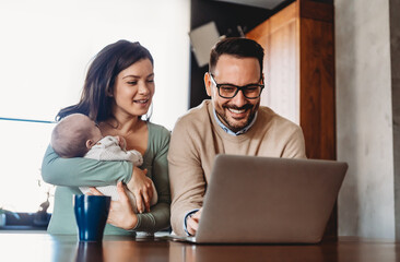 Portrait of young couple with a baby at home, using laptop. Father is trying to work from home.