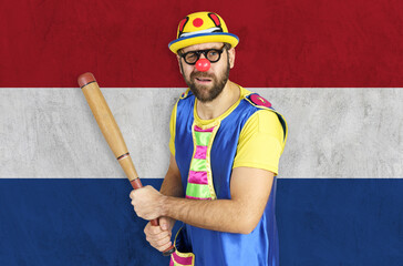 An insecure clown holds a bat in his hands against the background of the flag of Netherlands