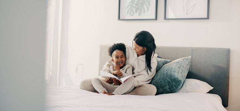 Mom and daughter laughing together as they read a story book on a bed