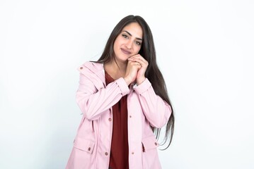 Charming serious young brunette woman wearing pink raincoat over white studio background  keeps hands near face smiles tenderly at camera