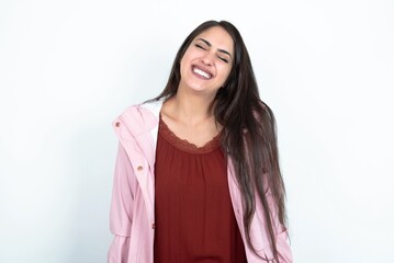 Fototapeta premium Positive young brunette woman wearing pink raincoat over white studio background with overjoyed expression closes eyes and laughs shows white perfect teeth