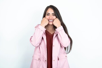 Happy young brunette woman wearing pink raincoat over white studio background  keeps fists on cheeks smiles broadly and has positive expression being in good mood
