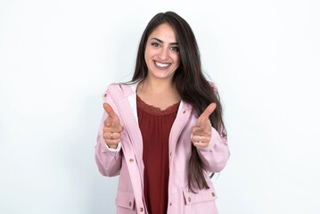 young brunette woman wearing pink raincoat over white studio background directs fingers at camera selects someone. I recommend you. Best choice