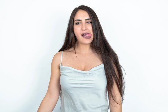 Funny young brunette woman wearing white tank top over white studio background makes grimace and crosses eyes plays fool has fun alone sticks out tongue.