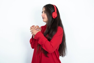 Young brunette woman wearing red shirt over white studio background wears stereo headphones listening to music concentrated and looking aside with interest.