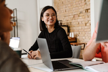 Asian woman smiling while sitting at table with her colleagues during meeting in office