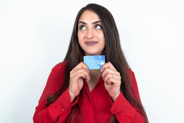 Photo of cheerful Young brunette woman wearing red shirt over white studio background hold debit card look empty space
