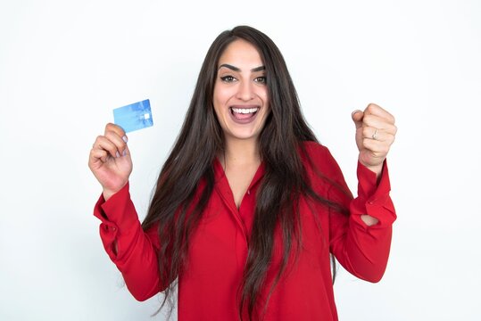 Photo of lucky impressed Young brunette woman wearing red shirt over white studio background arm fist holding credit card. Celebrated