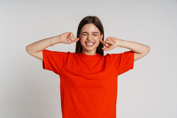Smiling woman covering her ears while standing with closed eyes isolated over white wall