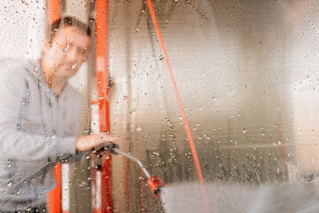 Man washing car with water gun in carwash self-service. Soap sud, wax and water drops covering vehicle window glass. Seen from the inside of the automobile.