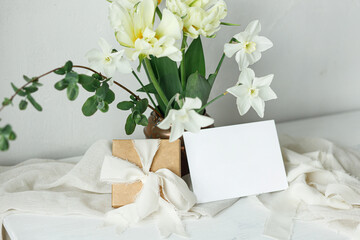 Beautiful bouquet in vintage vase, gift box and empty card on rustic wooden table. Greeting card mockup and stylish white spring tulips. Happy Mothers day. Women's day. Space for text