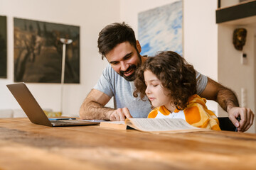 Father smiling while helping his son with homework sitting at table - obrazy, fototapety, plakaty