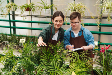 Man with down syndrome writing down notes while woman helping him to handle with flowers in greenhouse