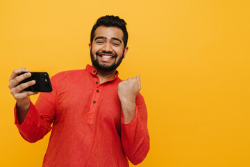 Indian man gesturing as winner while playing online game on mobile phone isolated over yellow...
