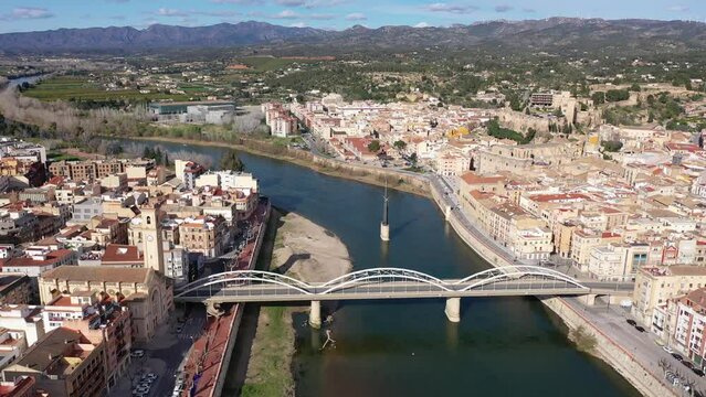 Aerial view of Catalan city of Tortosa on sunny spring day overlooking reconstructed Bridge of State across Ebro river and Monument to Battle of Ebro in middle of water, Spain