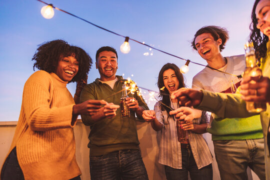 Multi-ethnic millennial group of friends folding sparklers on rooftop terrace at sunset, having beers, dinner with friends, New Years Eve, party,