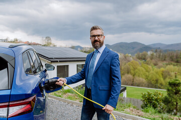 Mature businessman holding power supply cable and charging his electric car.