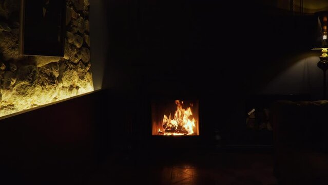 Natural logs burn intensely with flames in fireplace in dark room of rich aristocratic estate. Dark picture hangs on illuminated stone wall of family castle.