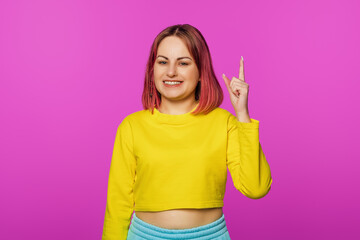 Portrait of pretty smiling young woman with pink hair looking at camera and pointing finger up, showing advertisement, company banner on top, pink background