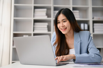 Smiling Asian businesswoman focusing on her business tasks on laptop, working in her office.