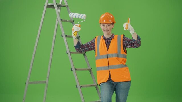 Green Screen.Young Happy Confident Female Builder in Checkered Shirt,Safety Jacket,Helmet,Gloves,Leaning on a Ladder,Lifting the Paint Roller Up,Making Thumbs Up and Smiling.Women at Work Concept