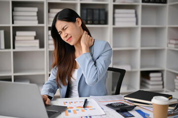 Overworked asian businesswoman sits at her desk rubbing her neck, suffering from neck pain