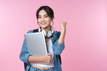 Portrait of smiling young Asian college student with laptop and backpack isolated over pink background.Cheer up success celebration concept