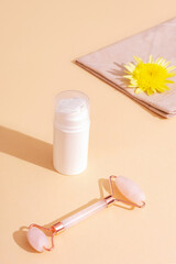 White cosmetic bottle, yellow flower and rose quartz facial roller on beige background in sunlight. Skincare, spa and wellness concept