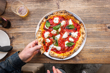 Male hand taking a slice of pizza freshly baked Neapolitan Margherita Pizza on a rustic wooden table in a traditional Pizzeria. - 595827406