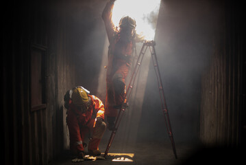 One firefighters checks the safety of the area while another attempts to climb a ladder in the galvanized room with soft light.