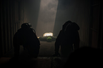 Two firefighters are sitting in the shadows and victims dummy lying on the floor in a galvanized room with light pouring in.