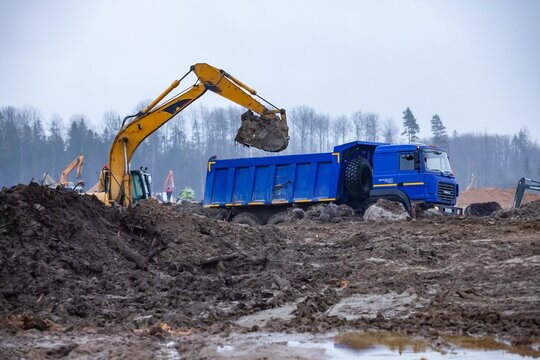 Ust-Luga, Leningrad oblast, Russia - November 16, 2021: Groundworks in rain, excavator loads dump truck. Flattening ground on site.