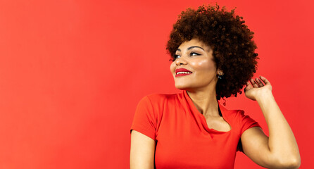 A dark-skinned smiling girl with curly afro hair posing on a red background in clothes of the same color. The young woman looks to the side while touching her hair. Copyspace.