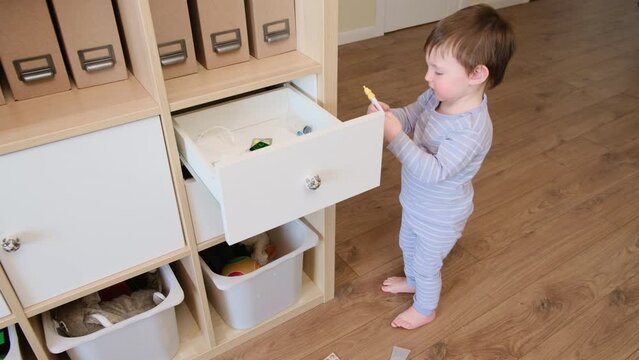 Toddler baby opened the cabinet drawer with pills and thermometer. Child boy holding a pack of pills in the home living room. Kid aged about two years (age one year nine months)