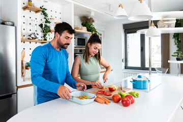 Happy young couple have fun in kitchen while preparing healthy organic food. Beautiful sports people are talking and smiling while cooking vegetarian meal in domestic kitchen at home.