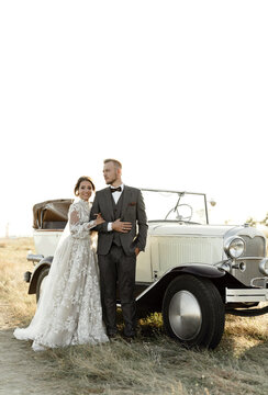 Young Groom Circles The Happy Bride At Sunset Of The Wedding Day Overlooking The Sea. Wedding Couple With Retro Car. Wedding Gift